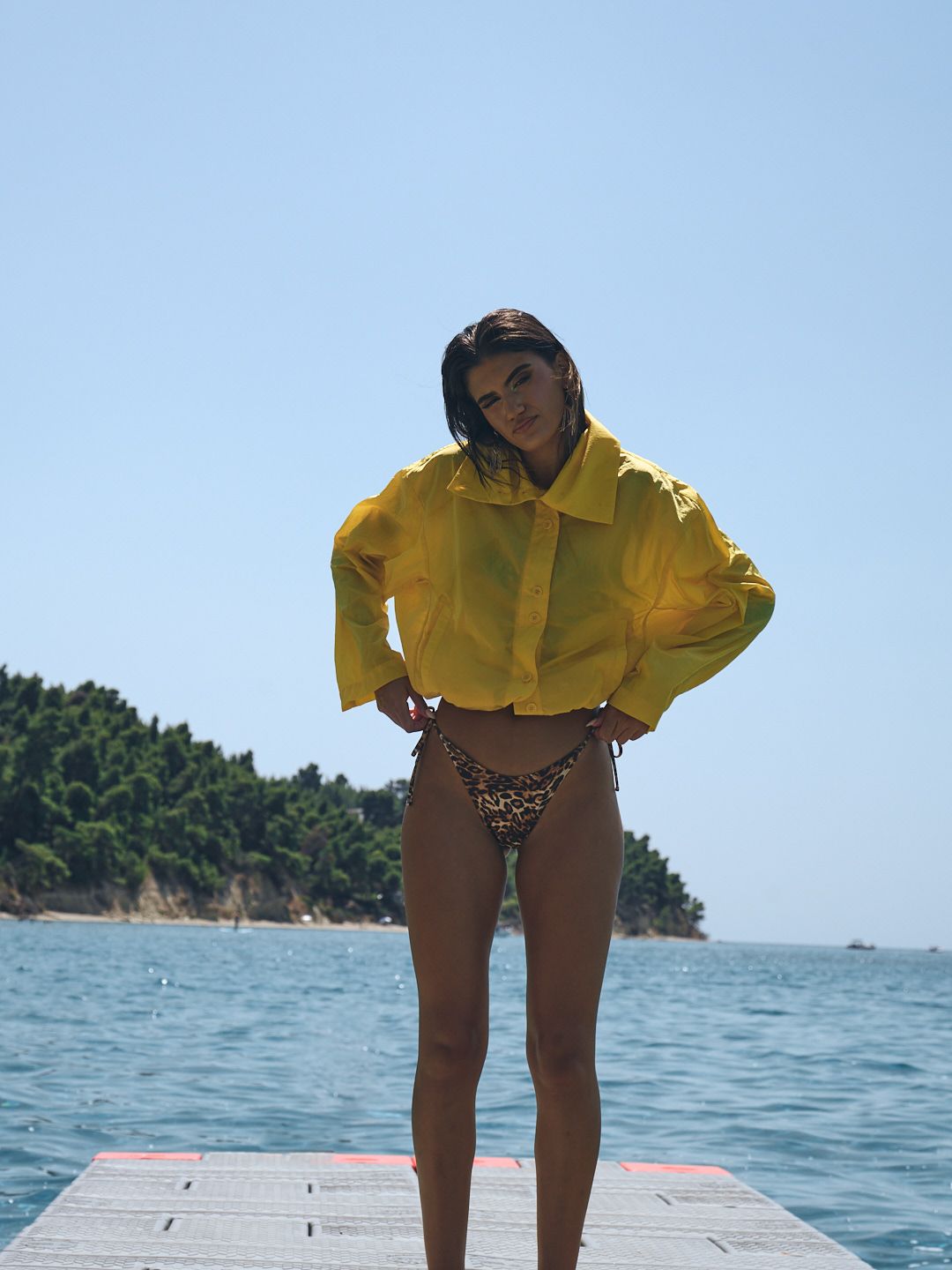 Woman in yellow jacket and leopard bikini bottoms posing on a dock over turquoise water on a sunny day.