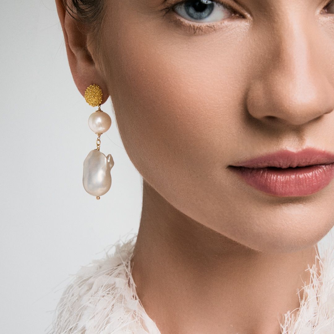 Woman wearing elegant gold and pearl drop earrings featuring a textured gold ball and baroque pearl pendant.