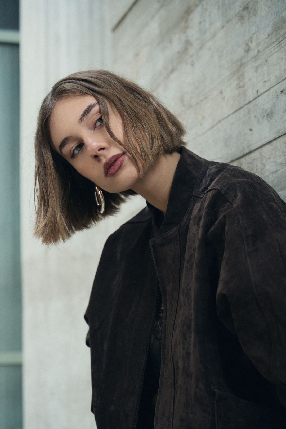 A fashion portrait of someone wearing a dark jacket and red lipstick against a white wall.