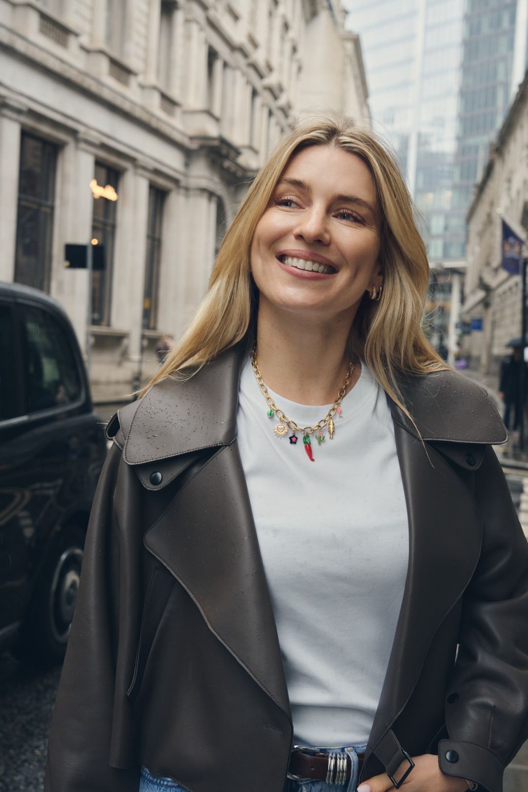 A person wearing a black leather jacket and white top smiles warmly while standing on an urban street.