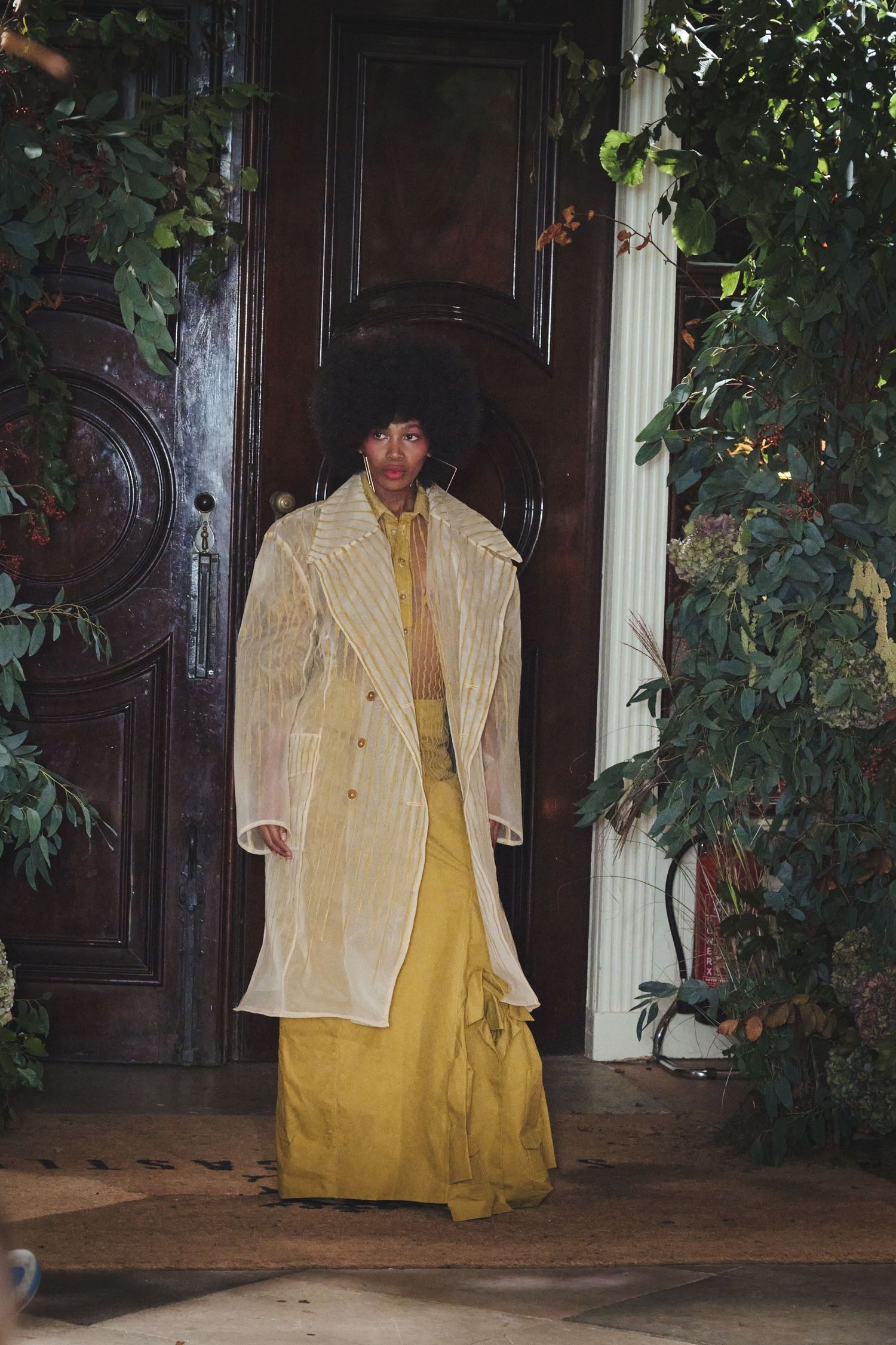 Fashion model walks runway in yellow dress with cream jacket during fashion show presentation.
