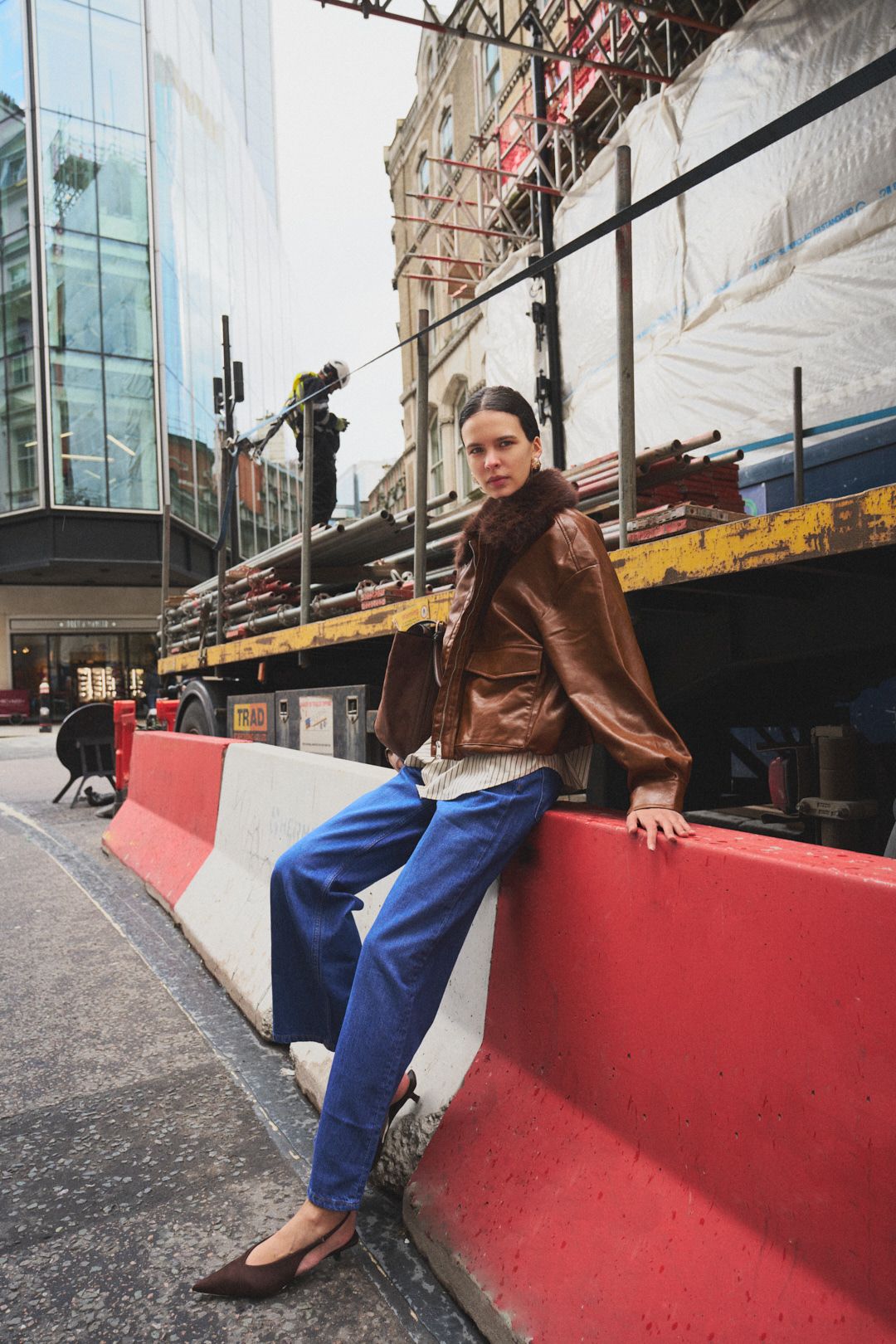 A stylish outfit featuring a brown leather jacket and blue jeans posed against a red construction barrier in an urban setting.