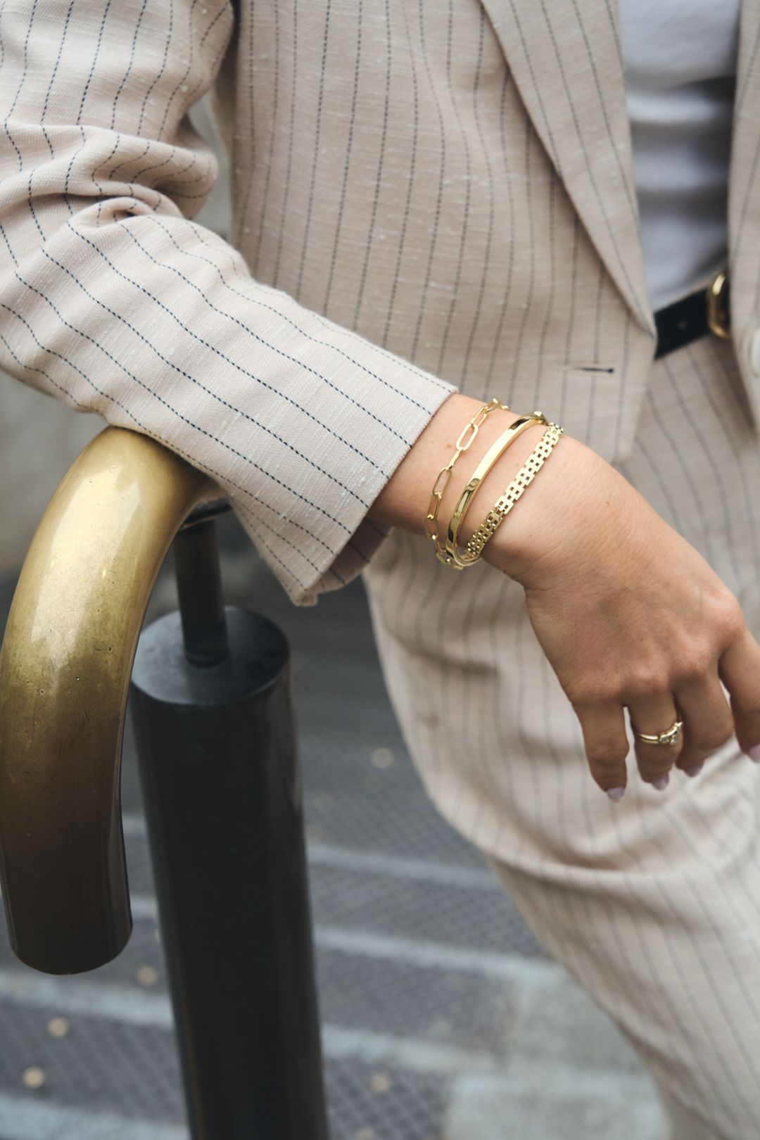 Elegant gold bracelets and rings accent a beige pinstriped suit sleeve against an urban railing.