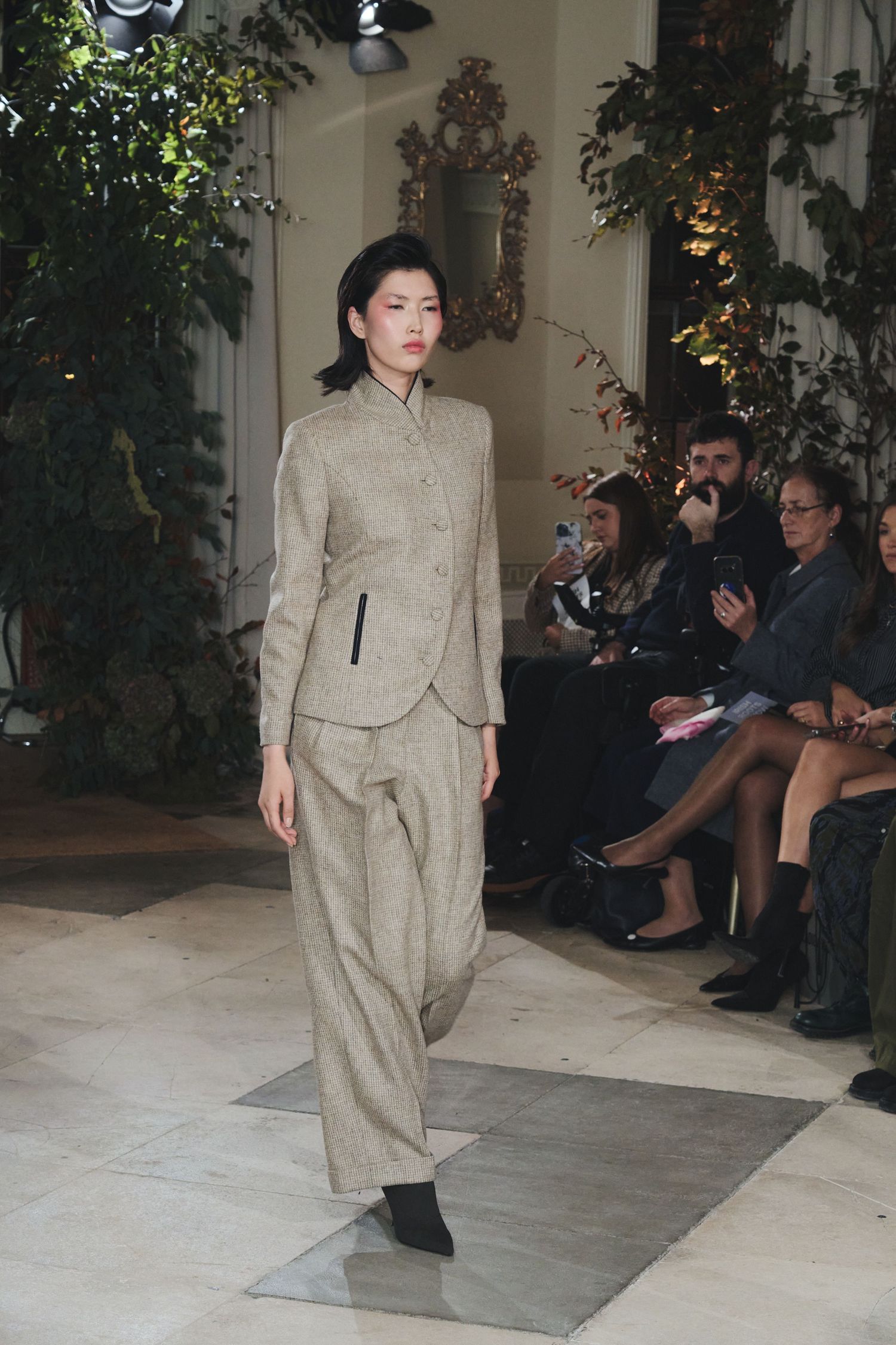 Model presents tailored beige linen suit ensemble on indoor runway with foliage backdrop.
