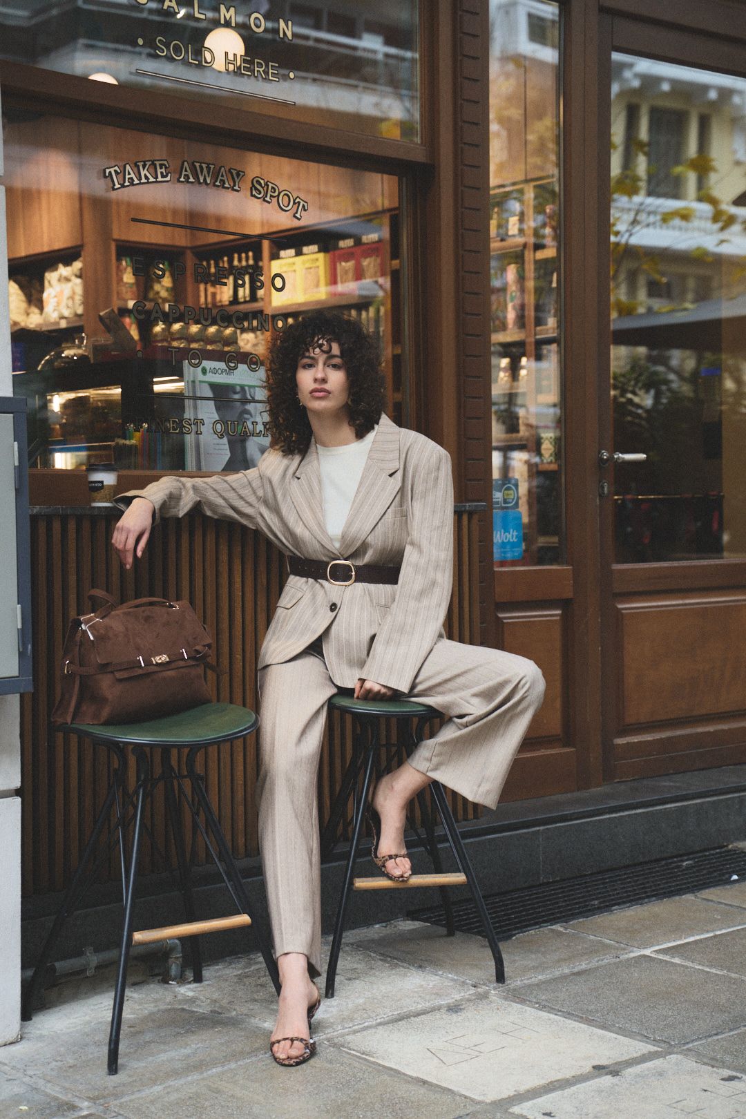 A person in a beige linen outfit sits on a bench outside a storefront with large windows.