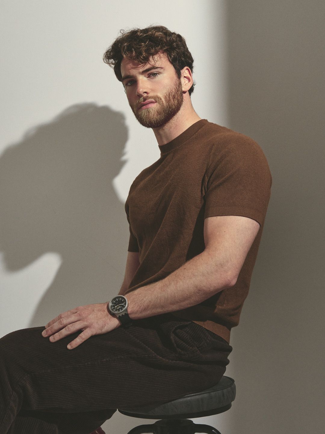 Fashionable model wearing brown t-shirt and dark pants sitting on chair against light backdrop with dramatic shadow.