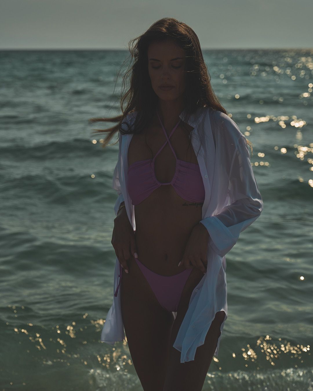 A figure wearing a purple bikini and white shirt stands against a sparkling ocean backdrop at sunset.