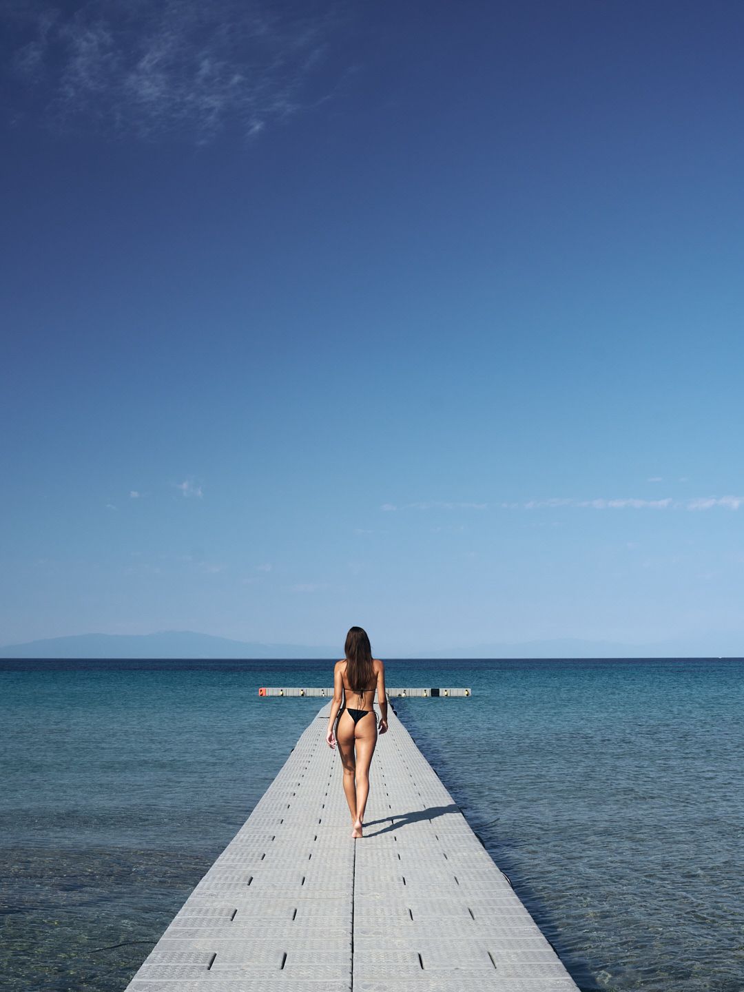 Woman in black bikini walking on a white pier toward the blue ocean under a clear sky.