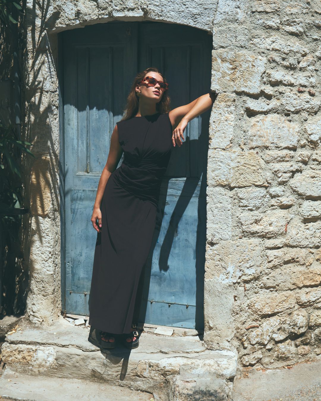 A series of four artistic photographs showing someone in black clothing posed against a rustic stone wall with blue doors.