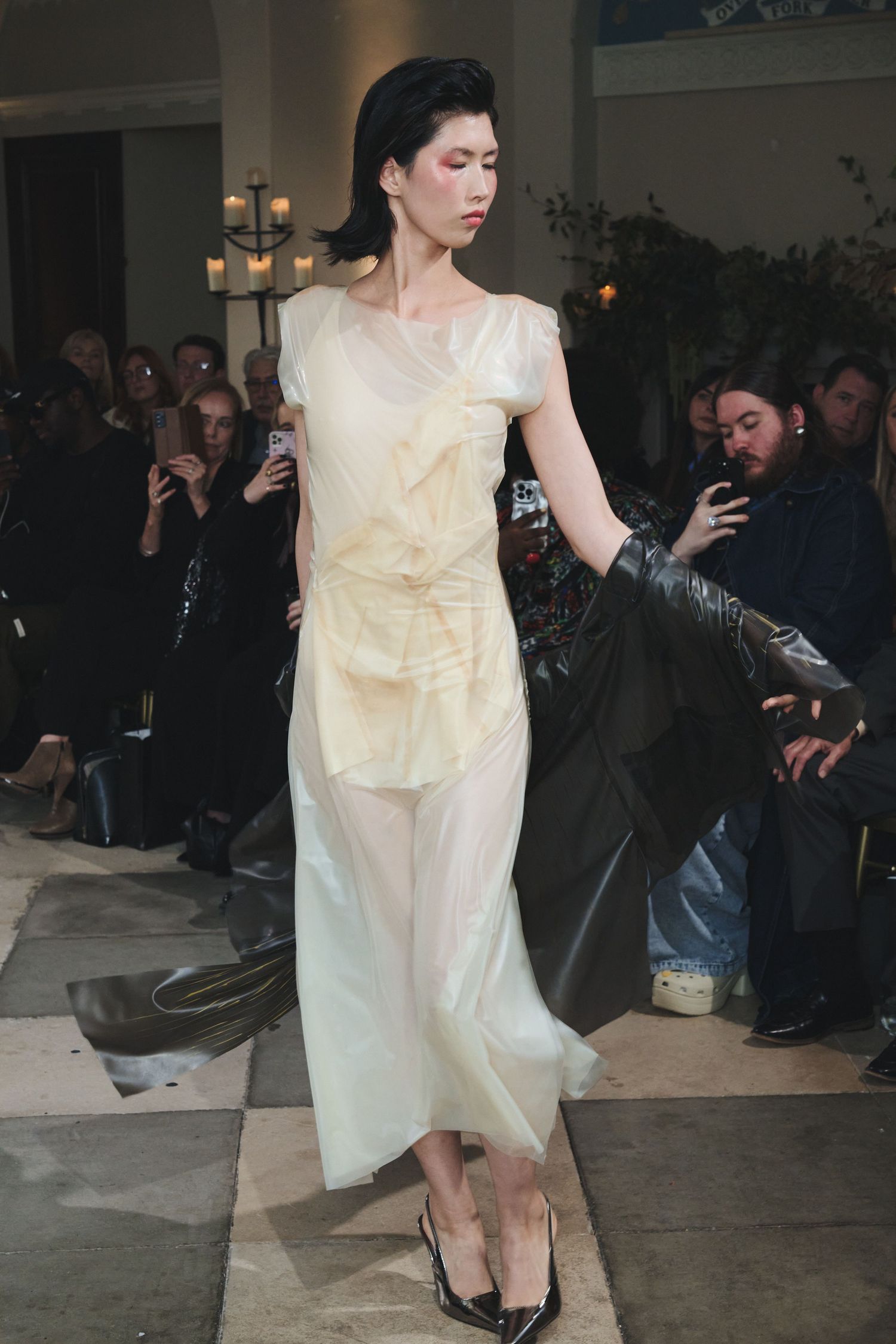 Fashion model in flowing cream dress performs dramatic pose on runway while audience watches in dimly lit venue.