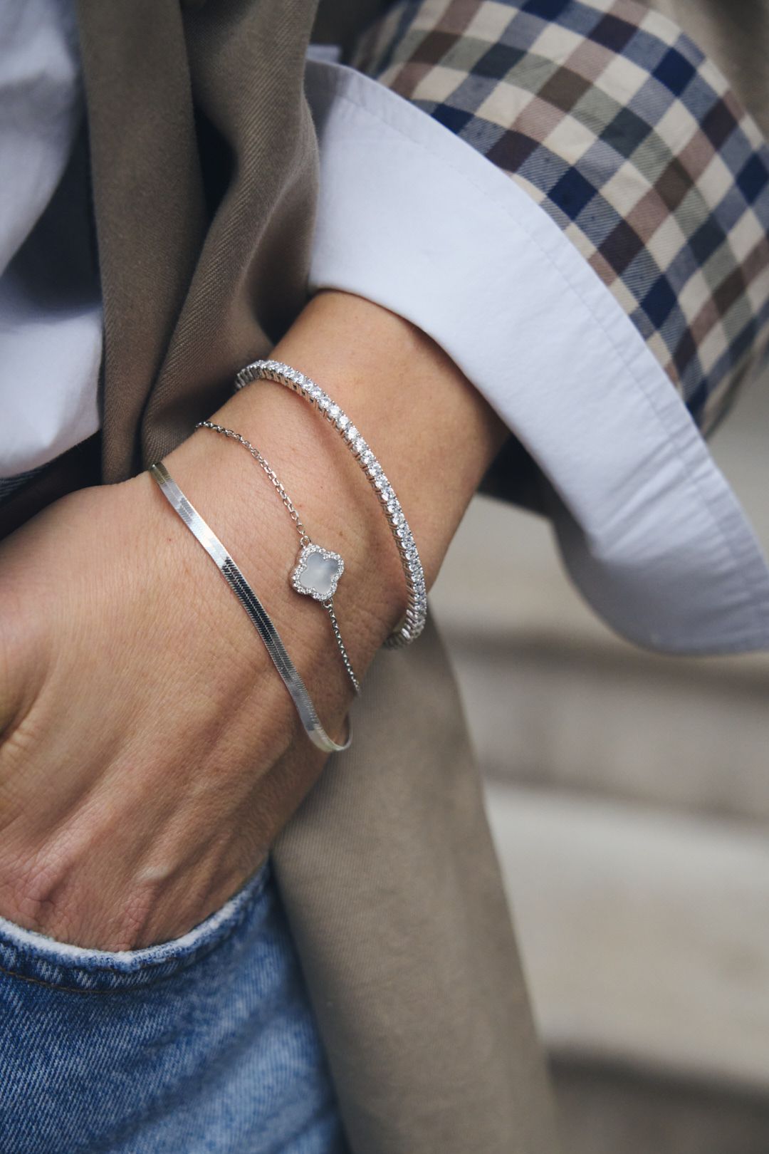 Multiple delicate silver bracelets and bangles styled together on a wrist with denim and plaid fabric visible.