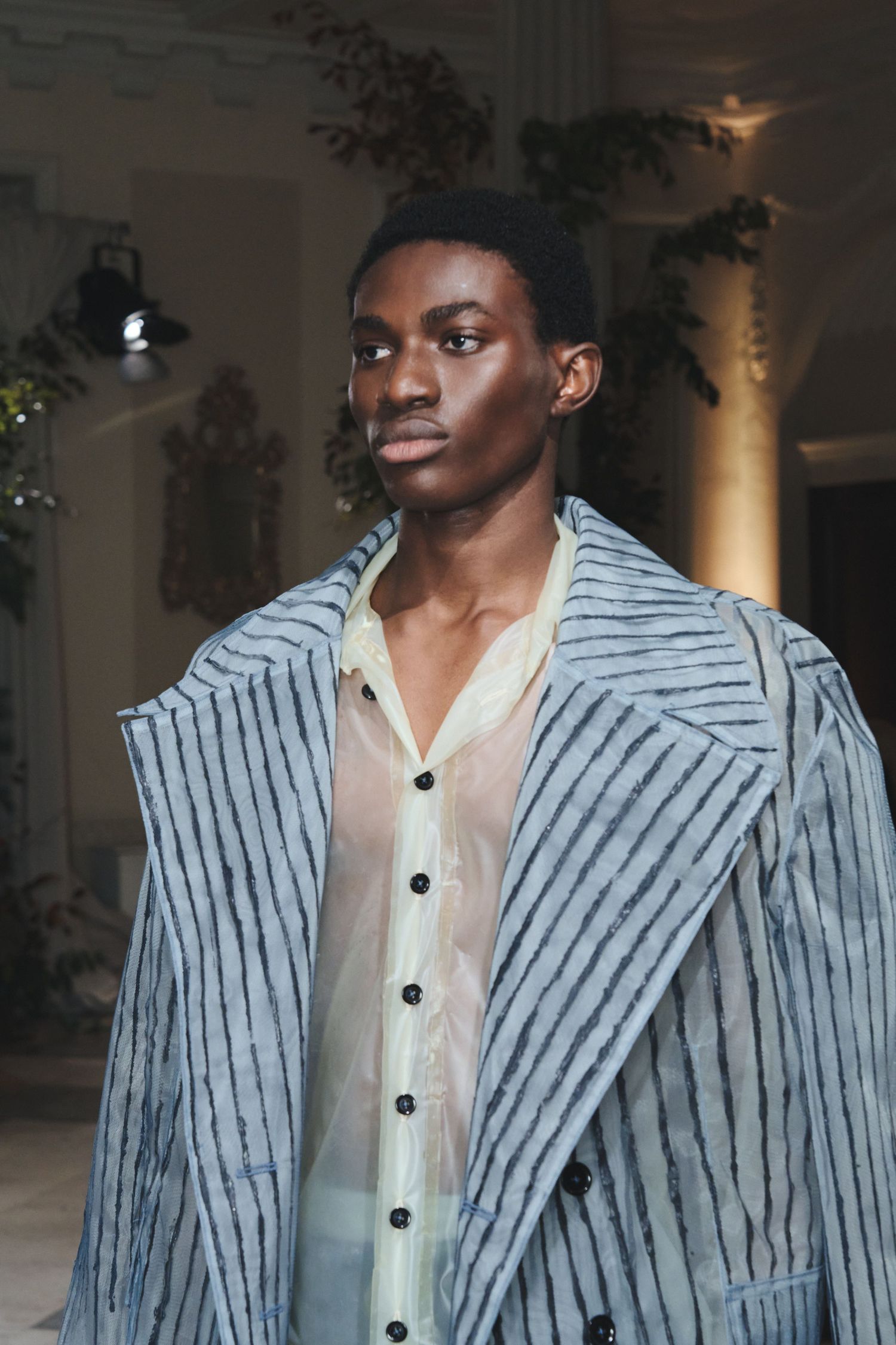 Person wearing a striped blazer and white button-up shirt poses in a dimly lit indoor setting.