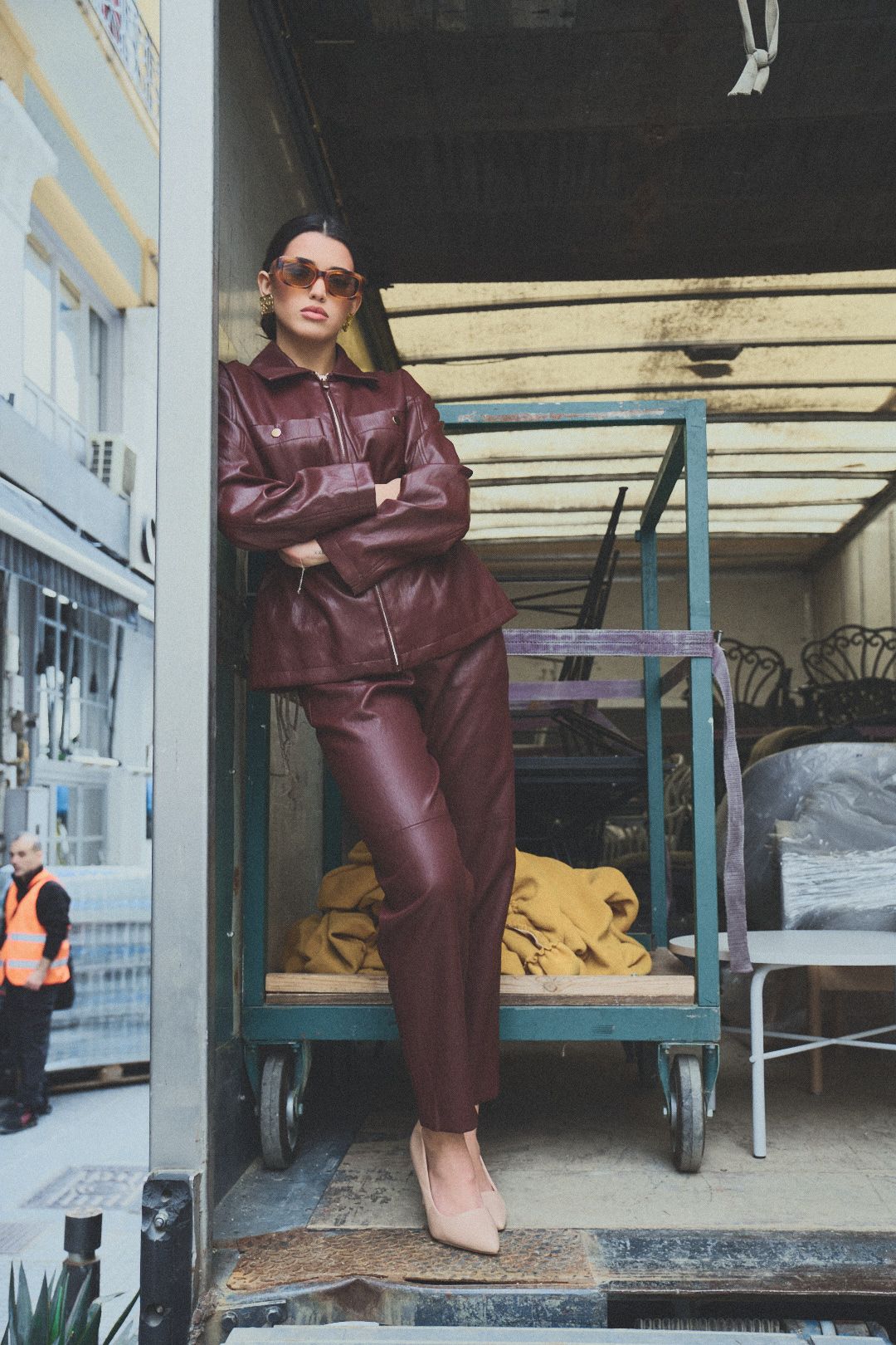 Woman in burgundy leather ensemble poses near industrial cart wearing sunglasses and beige heels.