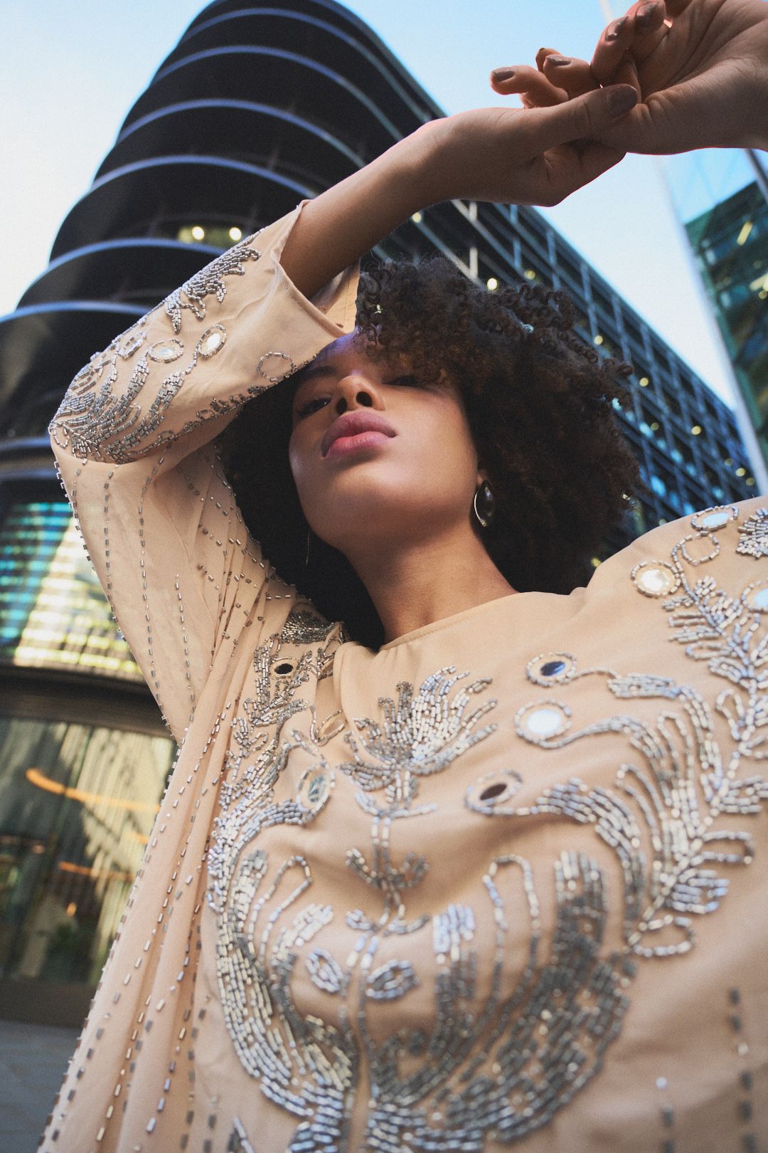 A person wearing a sheer beige dress with intricate silver beading and sequin embellishments poses dramatically outdoors.