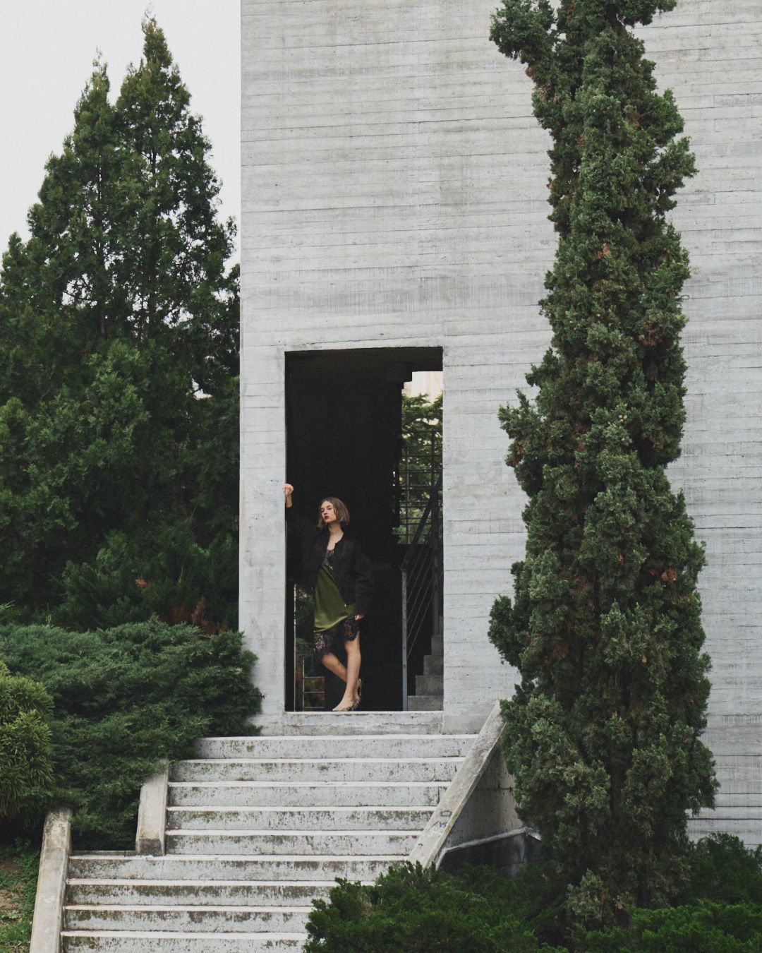 A figure stands in a doorway of a modern white building with concrete steps and tall cypress trees framing the entrance.