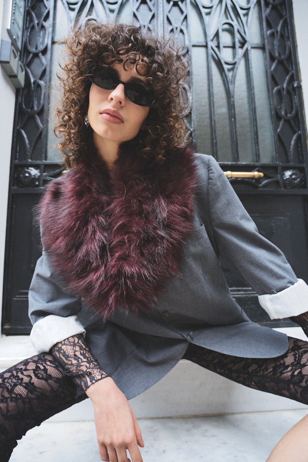 Fashion model in grey blazer with burgundy fur collar and lace sleeves poses against metallic background.