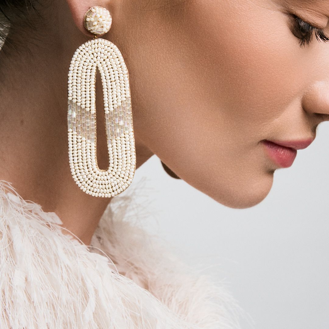 Woman wearing large oval crystal rhinestone drop earrings with pearl studs, styled with a white feather top.