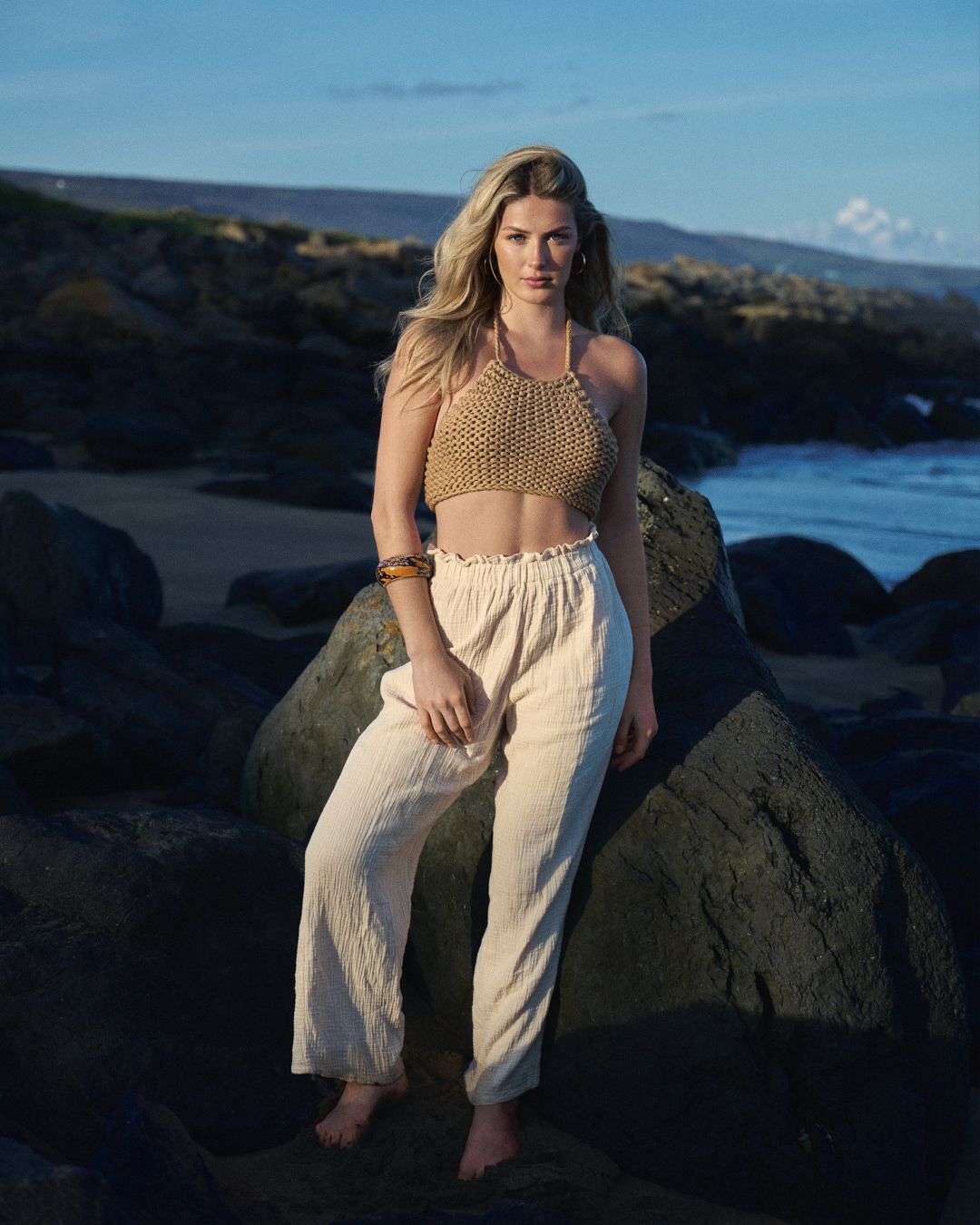 Model wearing crochet crop top and linen pants poses on rocky beach during golden hour.