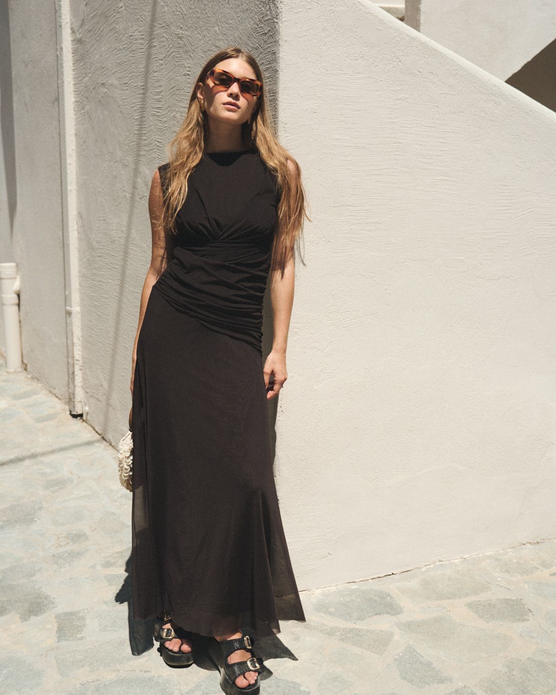 A model in sunglasses poses outdoors in a long black maxi dress against a white wall on a sunny day.
