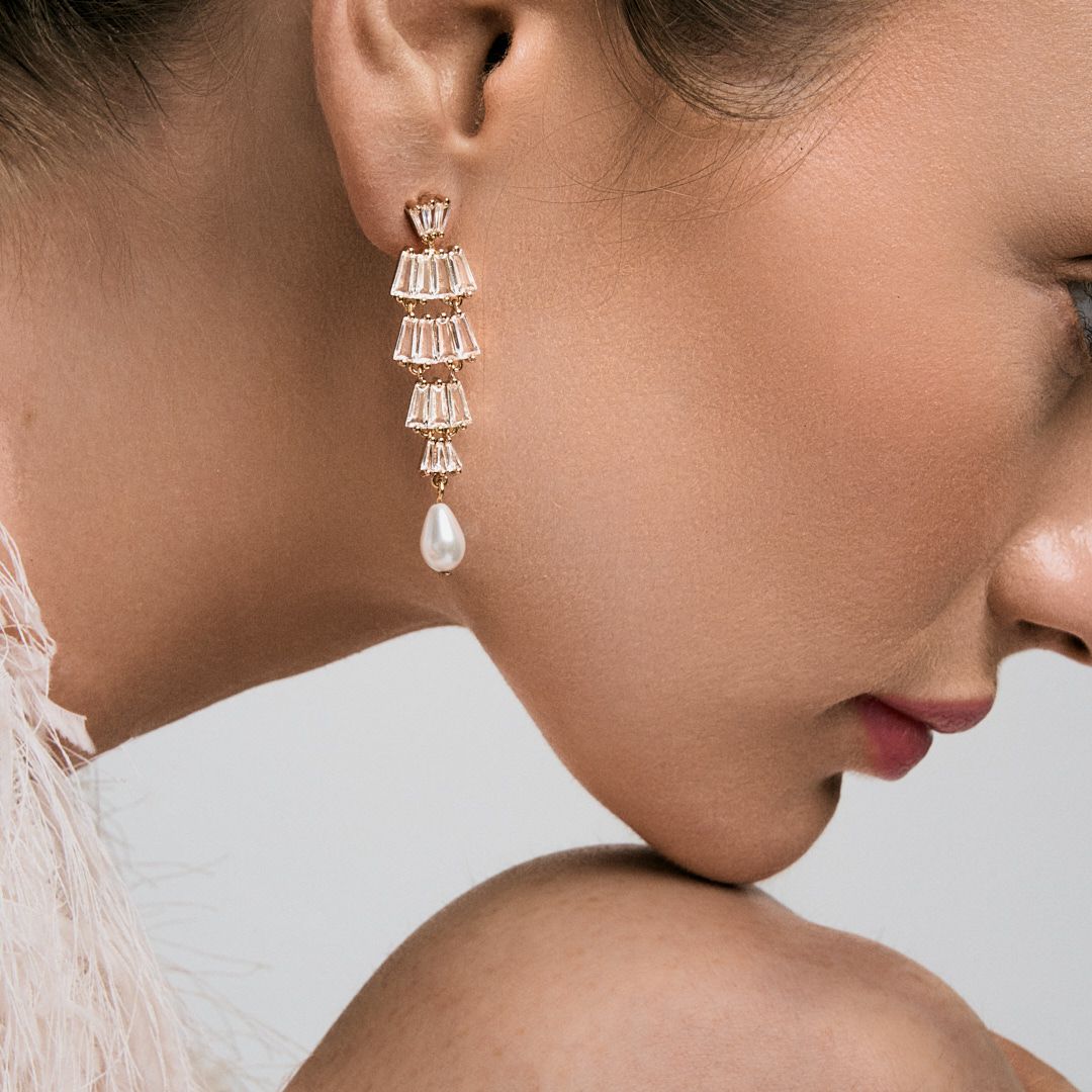 Gold drop earrings featuring cascading baguette crystals and a pearl pendant, worn by a model with elegant styling.