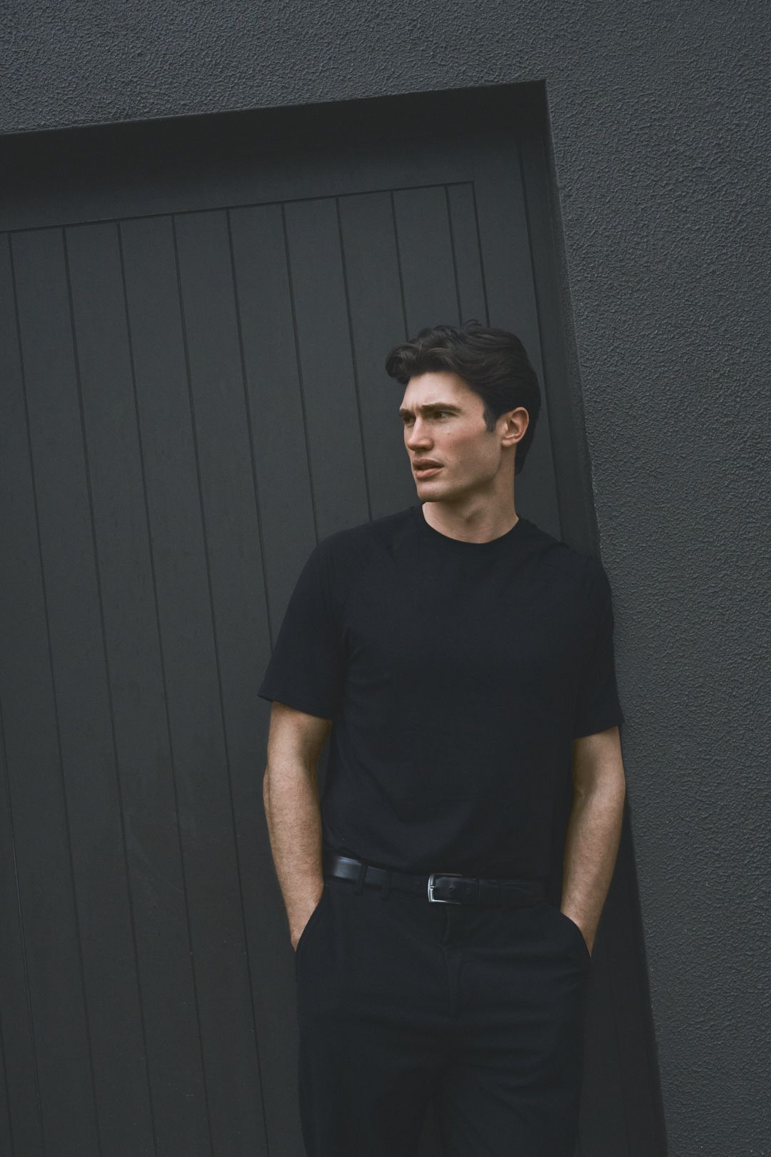 A person in a black t-shirt poses against a dark grey wall in a series of moody fashion portraits.