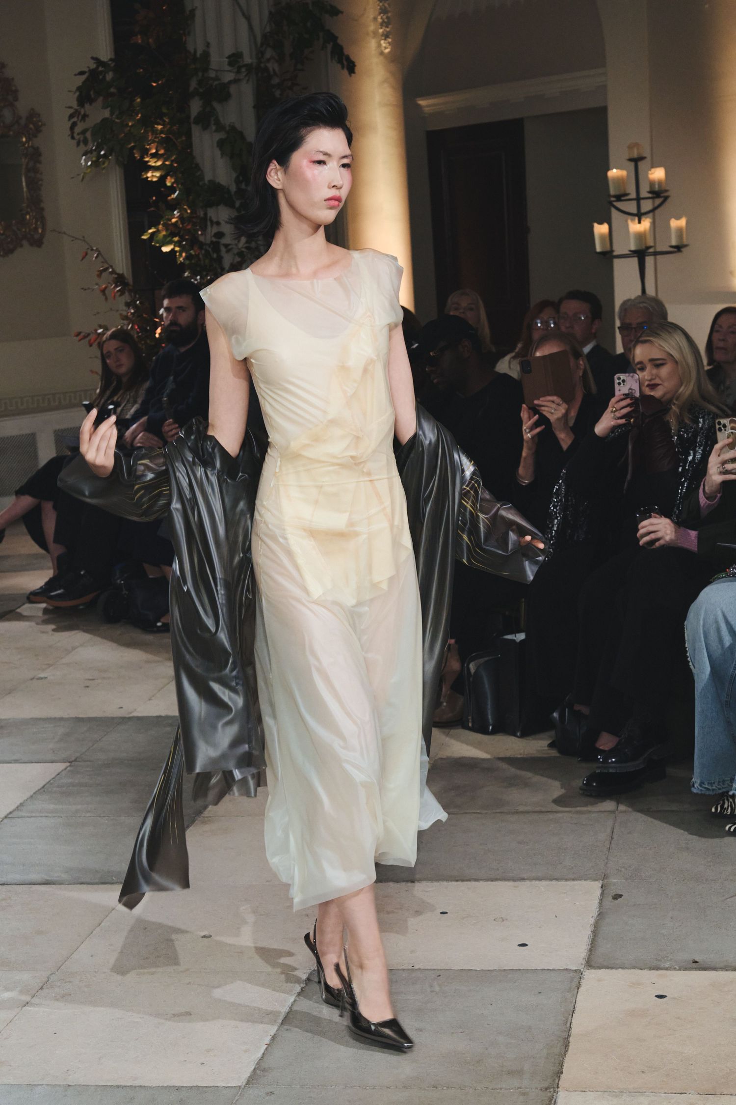 Model showcases flowing cream-colored evening dress with elegant draping on fashion runway.