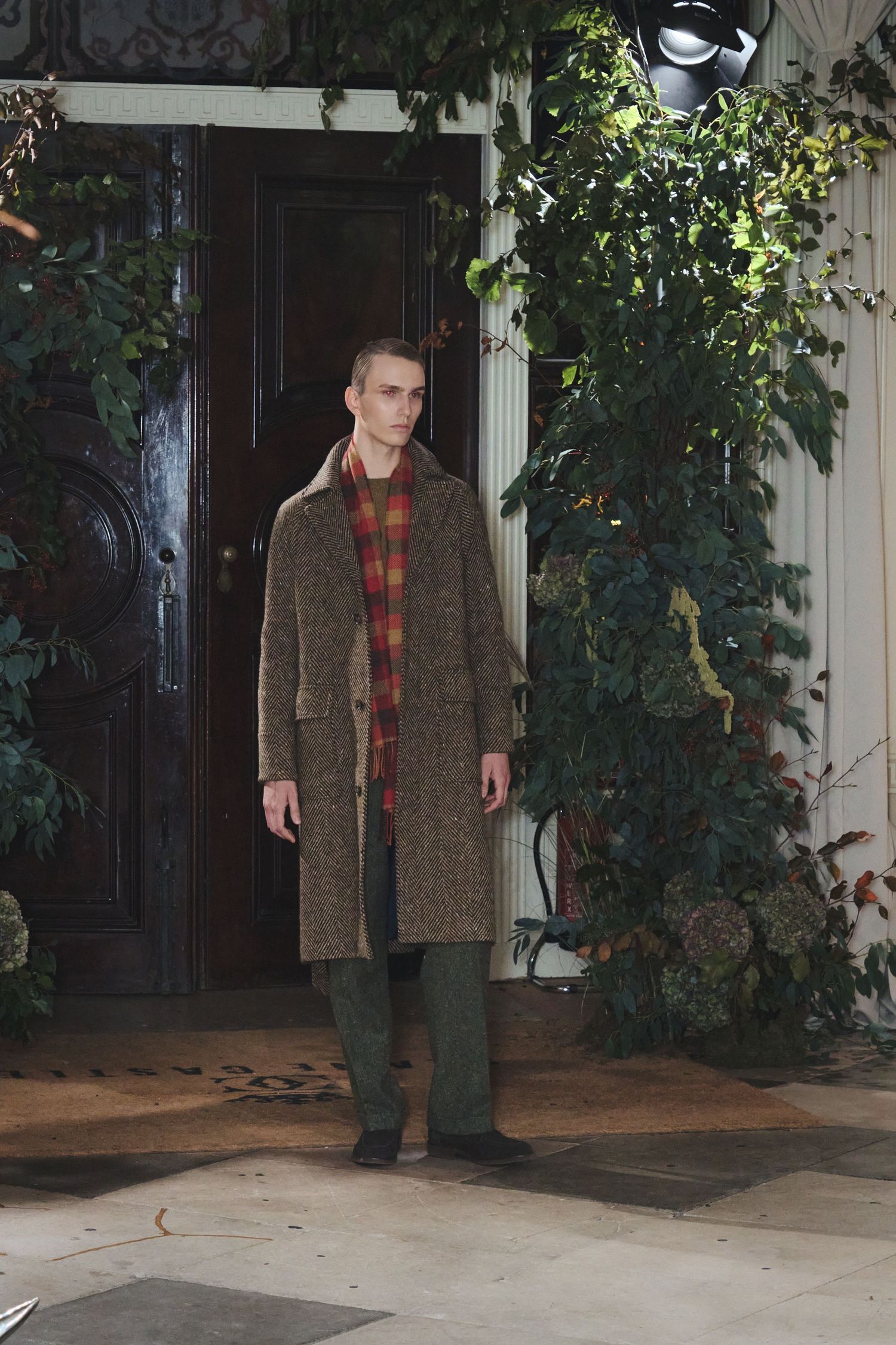 A person wearing a brown tweed coat and plaid shirt stands in front of a door with climbing vines on the wall.