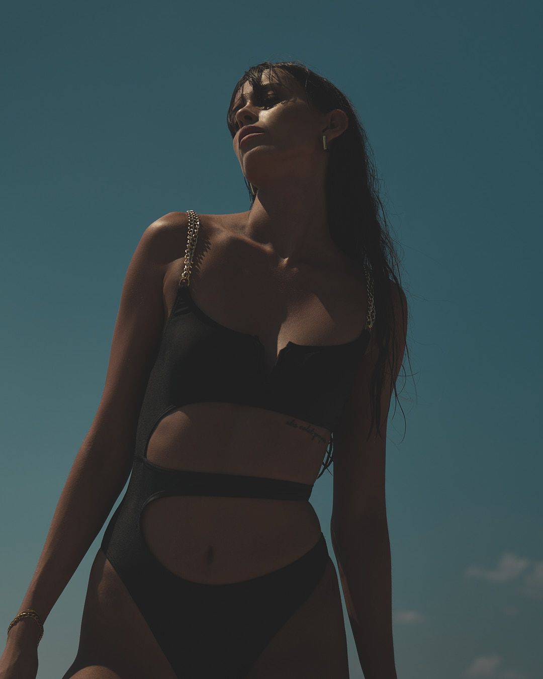 Silhouetted figure in black bikini poses against blue sky in dramatic low angle beach photoshoot.