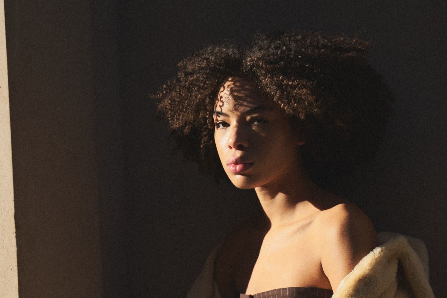 A person with curly hair stands in dramatic lighting against a dark background, creating a striking silhouette effect.