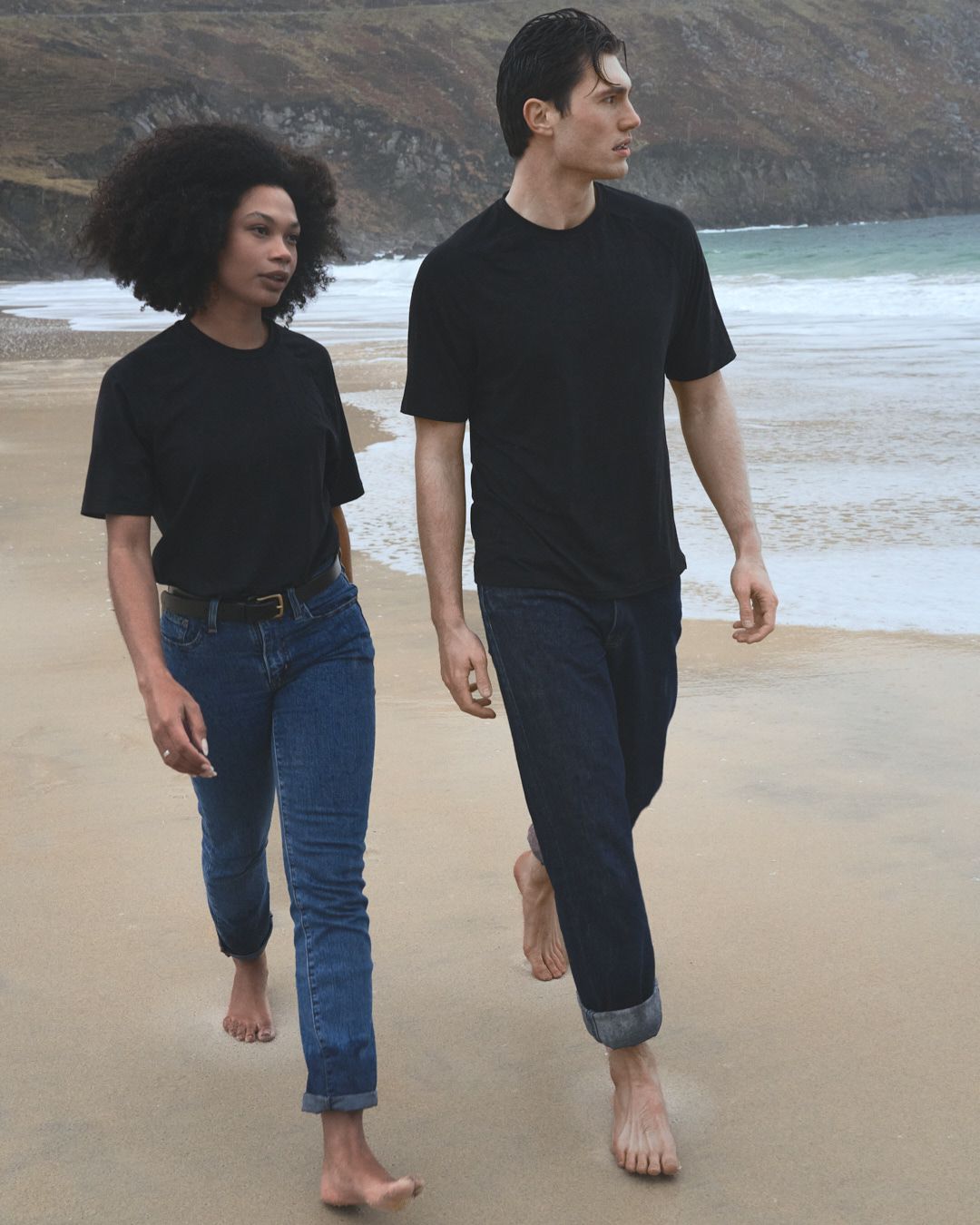 Two people walking barefoot on a beach wearing black t-shirts and jeans with coastal cliffs in the background.