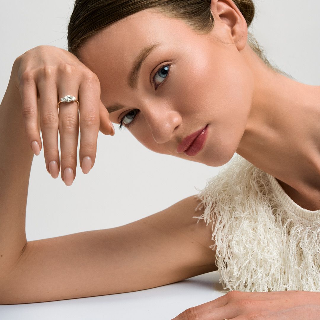 A woman wearing a white feathered top and a diamond ring poses elegantly against a light gray background.
