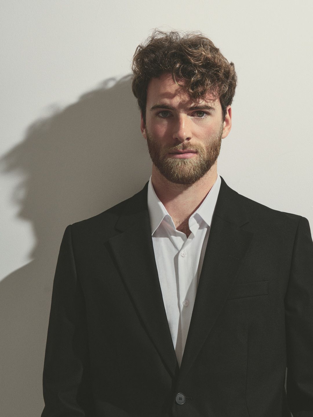 Professional portrait photograph of a person in a black suit and white shirt against a neutral background with dramatic shadow.