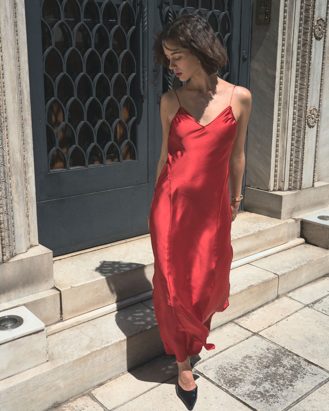 Woman in a flowing red dress stands elegantly against a stone building with ornate architectural details.