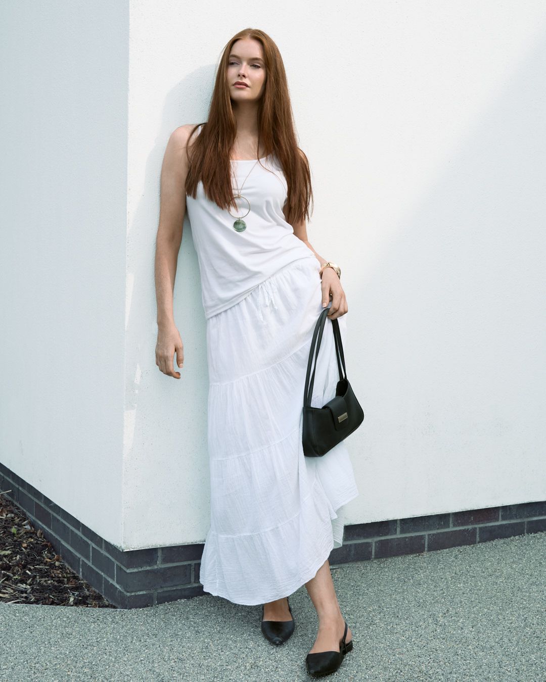 A person in a flowing white maxi dress and ballet flats poses against a white wall while holding a black handbag.