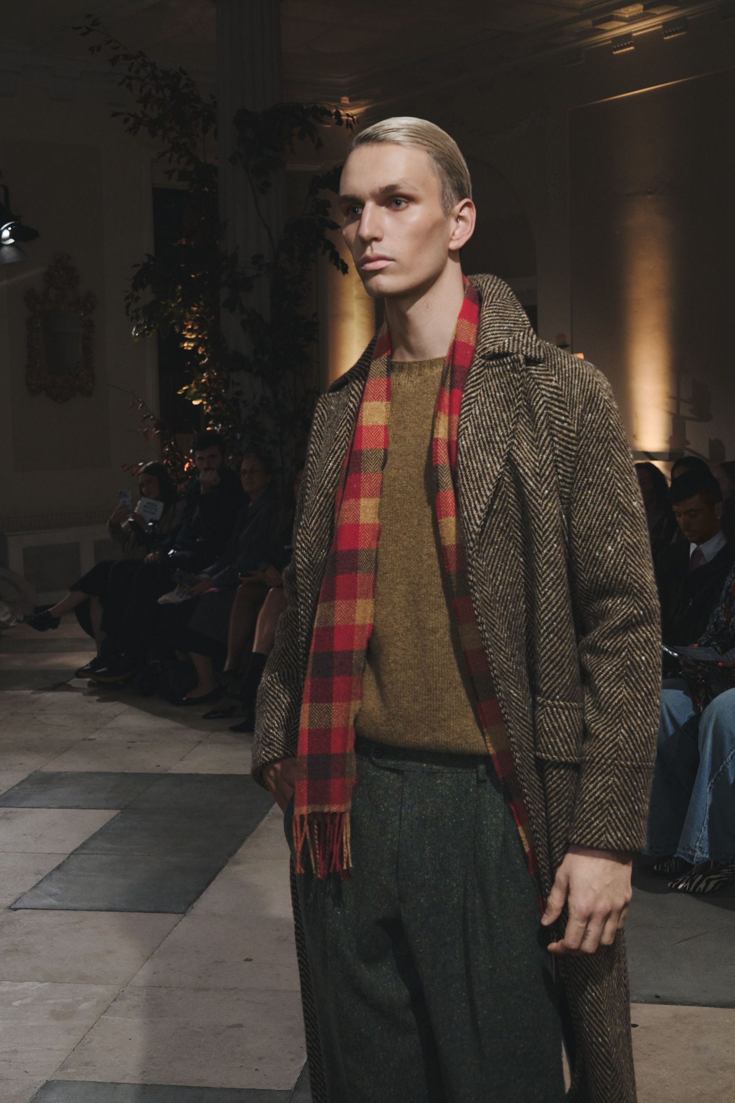 Runway model wears tweed coat with plaid scarf and dark trousers in autumn collection presentation.