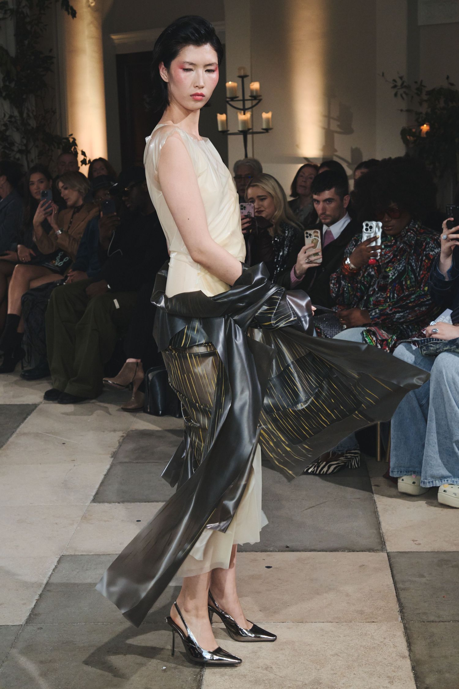 Model demonstrates dramatic movement in cream dress and black coat during fashion show performance.