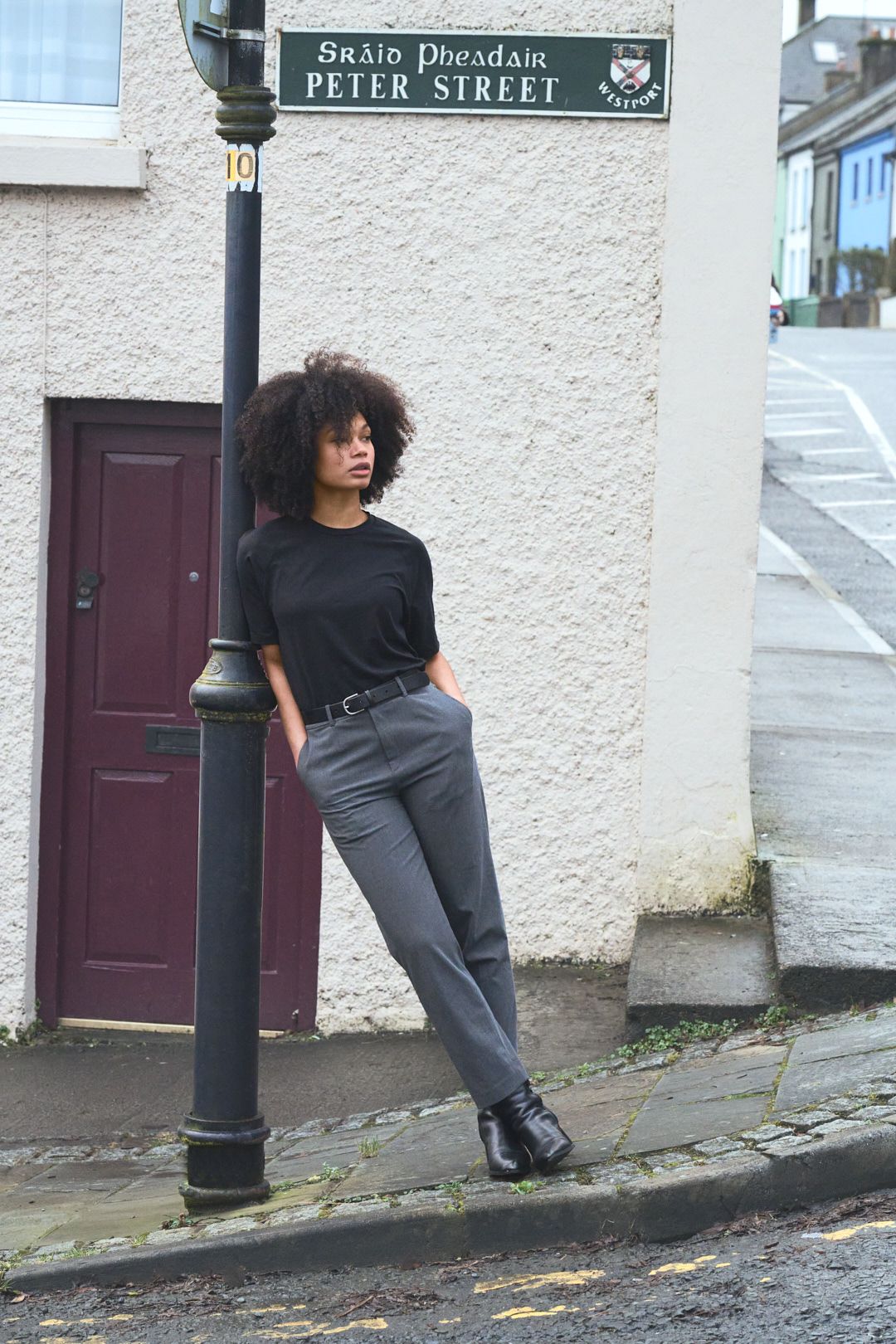 A person in black and grey clothing poses dramatically against a white wall near a street sign and door.