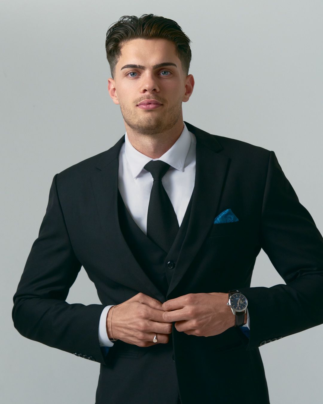 Professional portrait of someone wearing a black three-piece suit with blue pocket square and wristwatch.