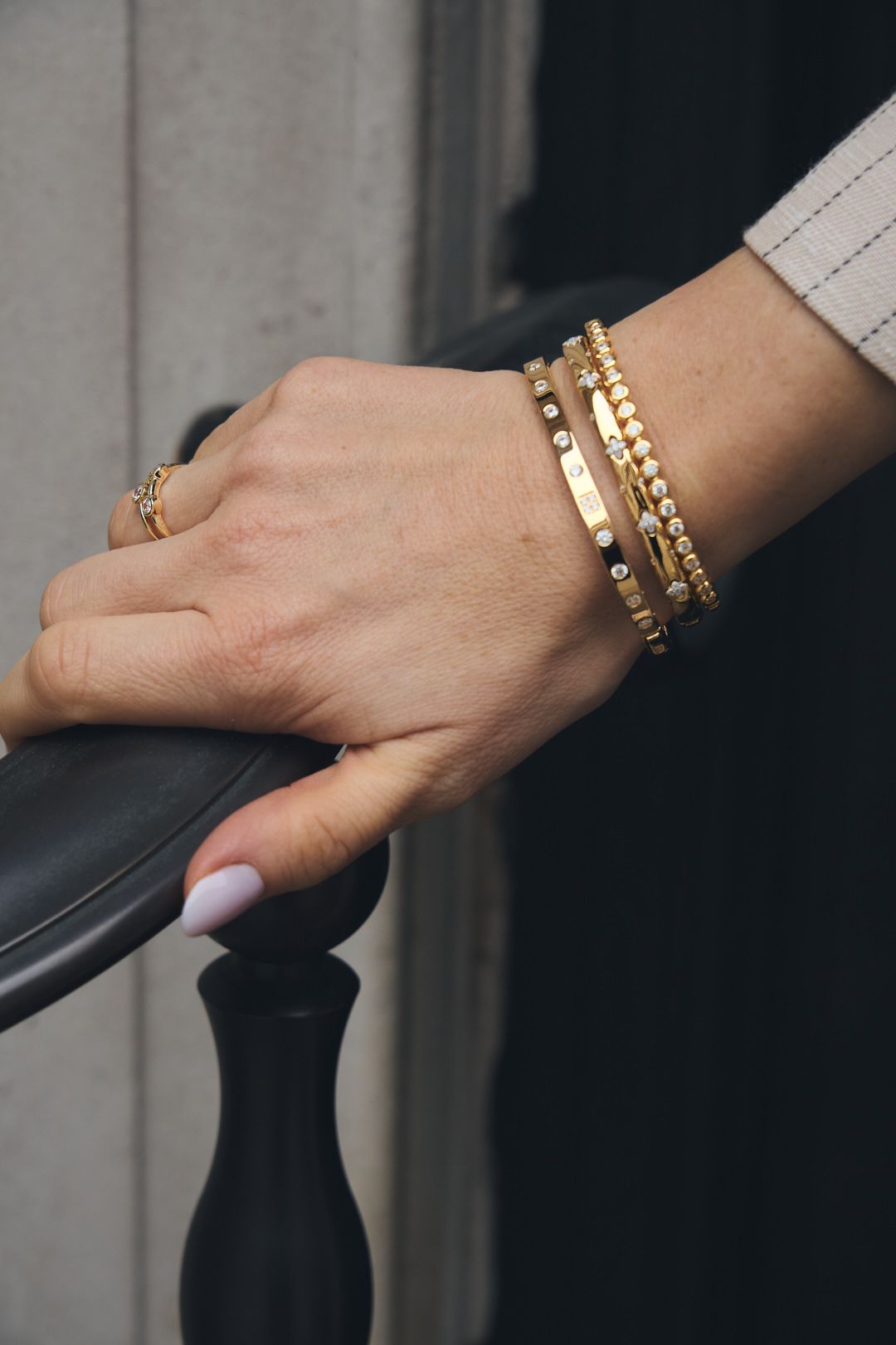 Close-up of a hand wearing delicate gold chain bracelets and rings with a black railing in the background.