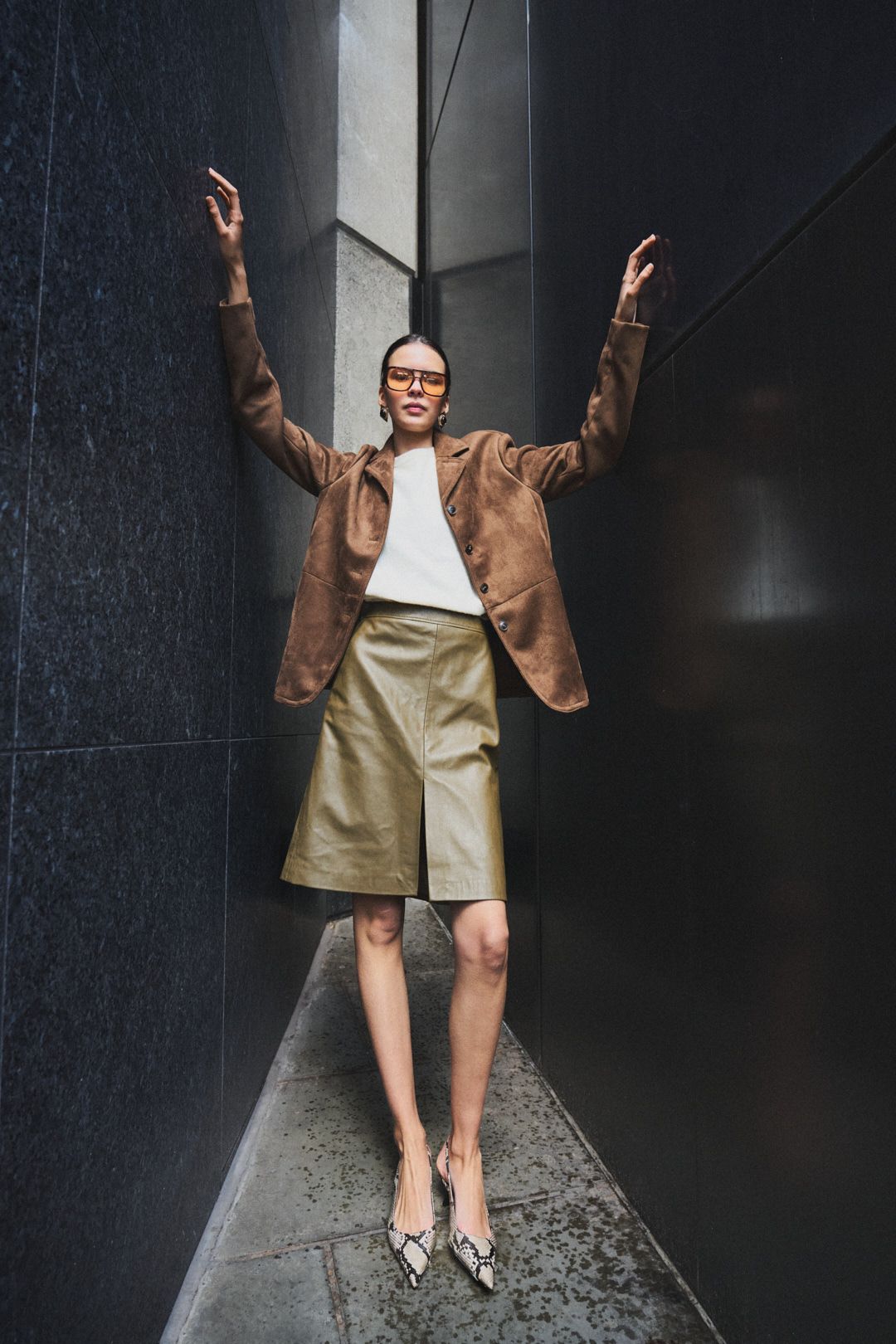 A person in a brown blazer and beige shorts poses in a dark hallway with a textured wall.