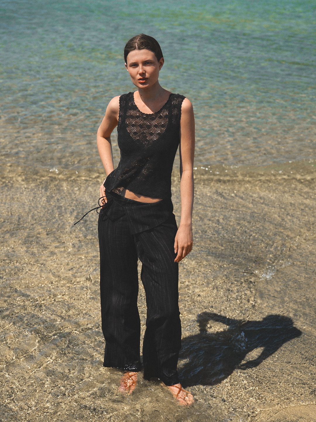 Woman in black lace top and wide-leg pants standing barefoot in shallow ocean water on a sunny beach.