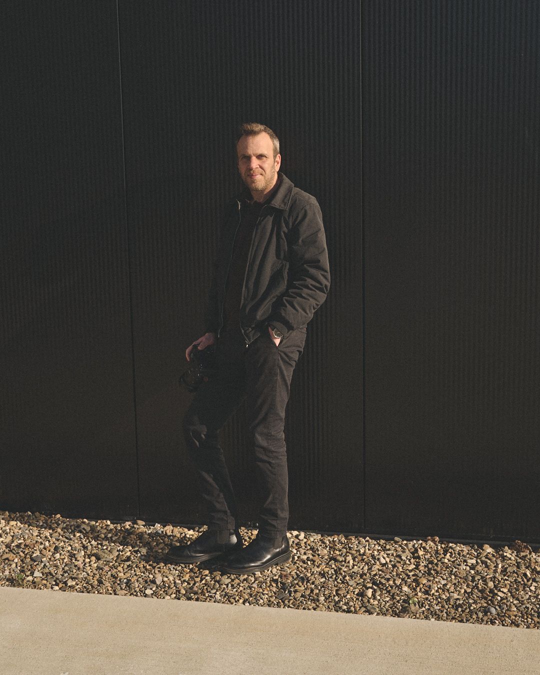 A person wearing dark clothing stands casually against a black wall with gravel at their feet in moody lighting.