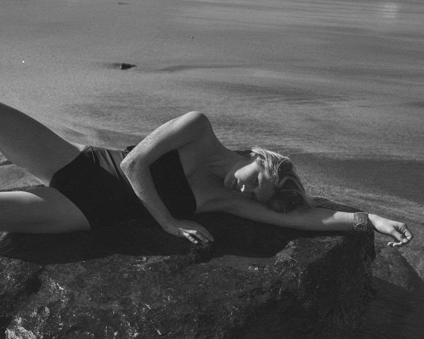 A figure in black swimwear reclines on dark rocks by the ocean.