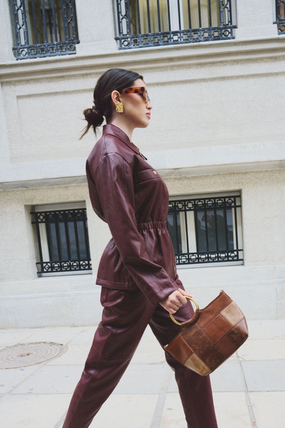 Stylish woman in burgundy leather outfit walks confidently on city street while carrying brown handbag.