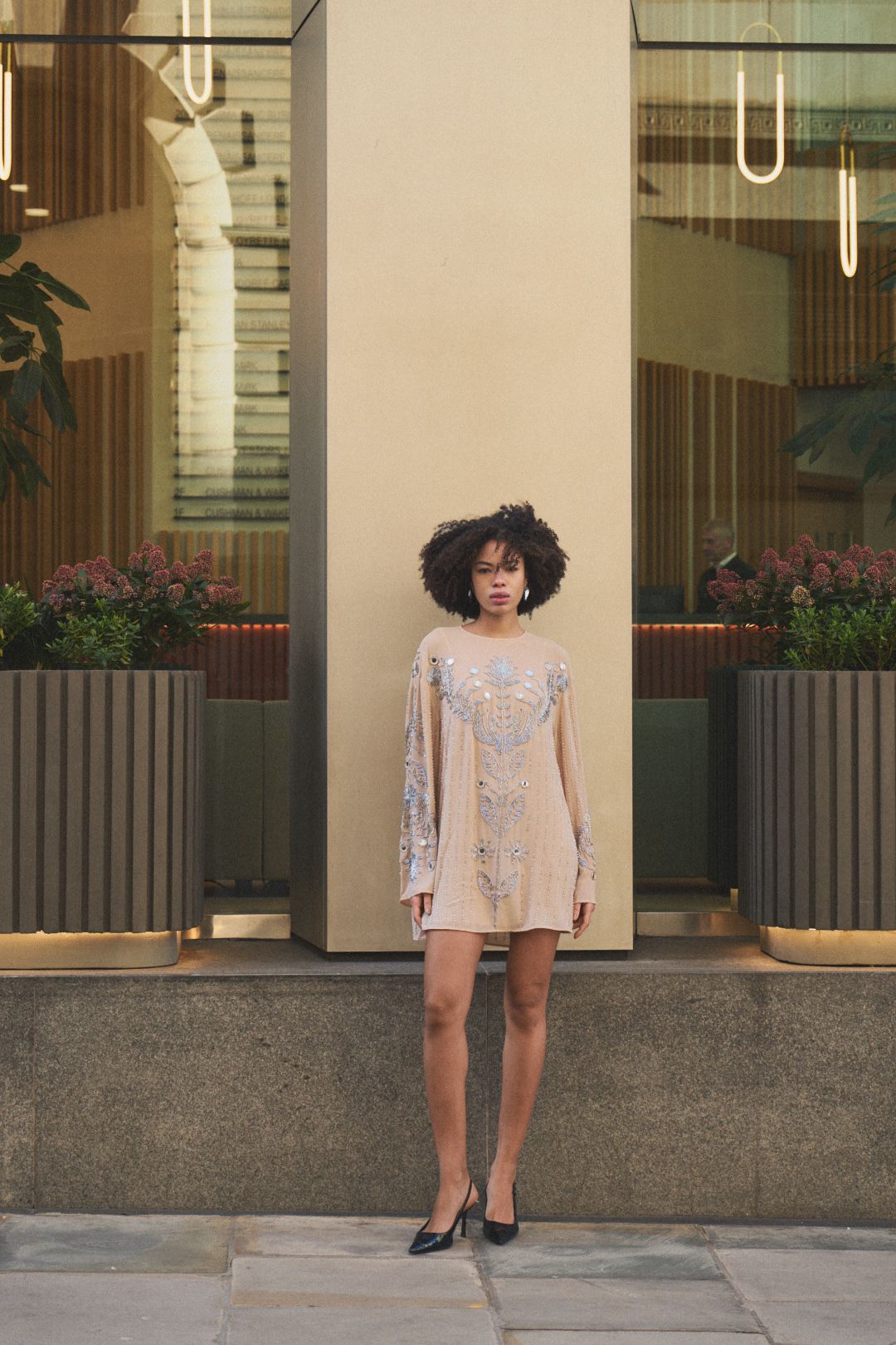 A person in a light lace dress poses against a modern building exterior.