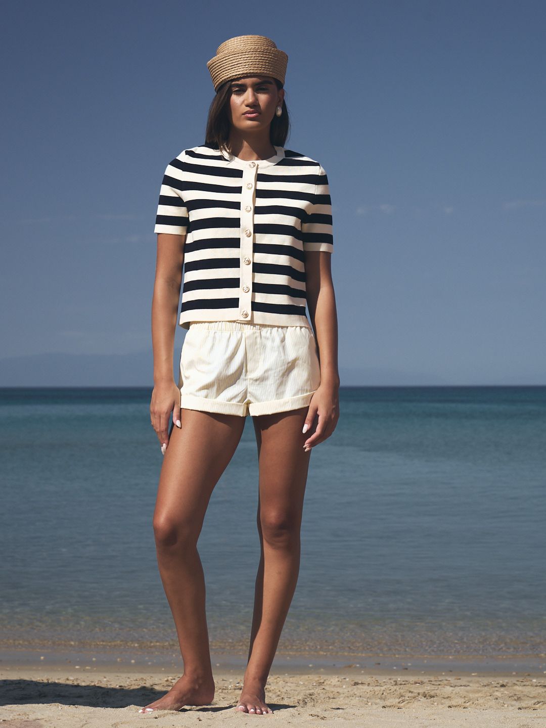 Woman modeling navy striped knit top with cream shorts and straw cap standing on a sunny beach.