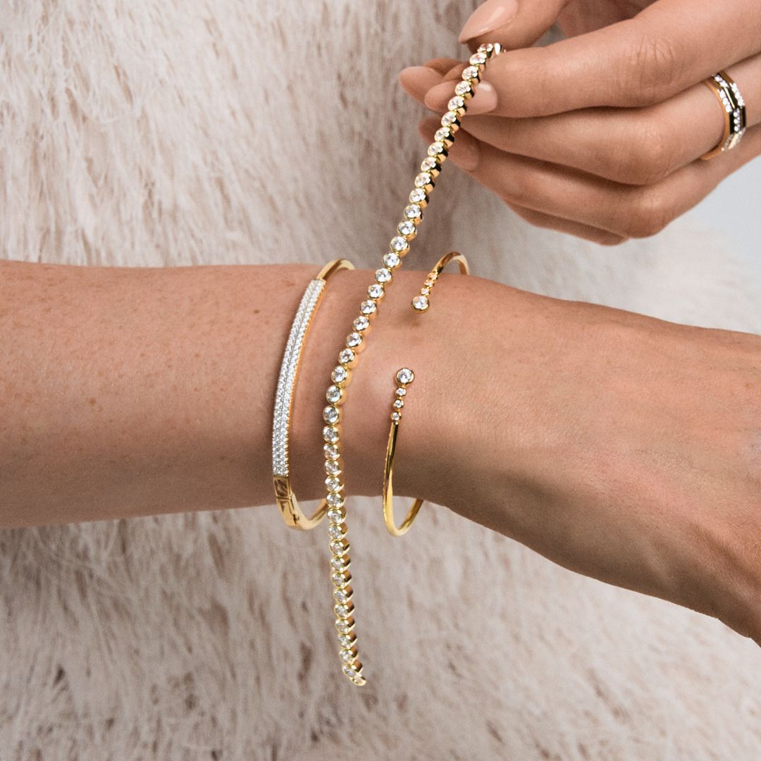 Woman wearing stacked gold bangles and bracelets with diamonds on wrist, holding a delicate pearl bracelet.