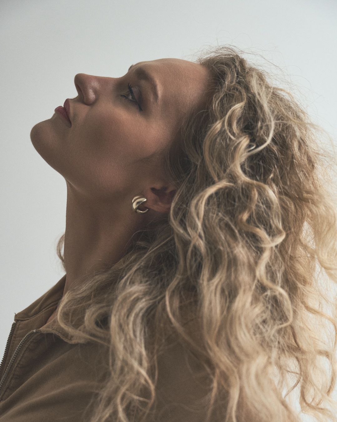 Profile view of a person with long curly blonde hair tilting their head back while wearing a brown jacket.