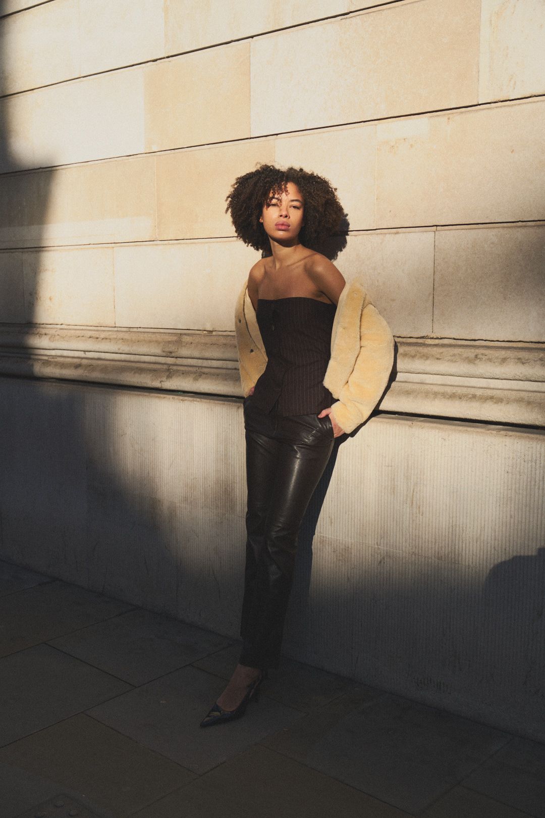A person in a beige coat and black pants poses against a light-colored wall in dramatic lighting.