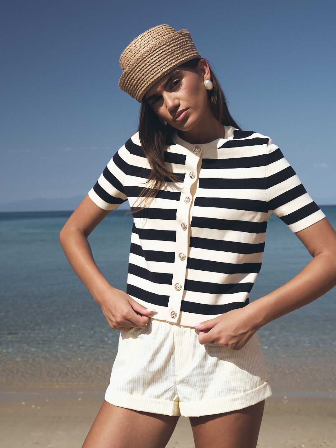 Woman wearing navy striped knit cardigan, white shorts, and woven cap posing on a sunny beach.